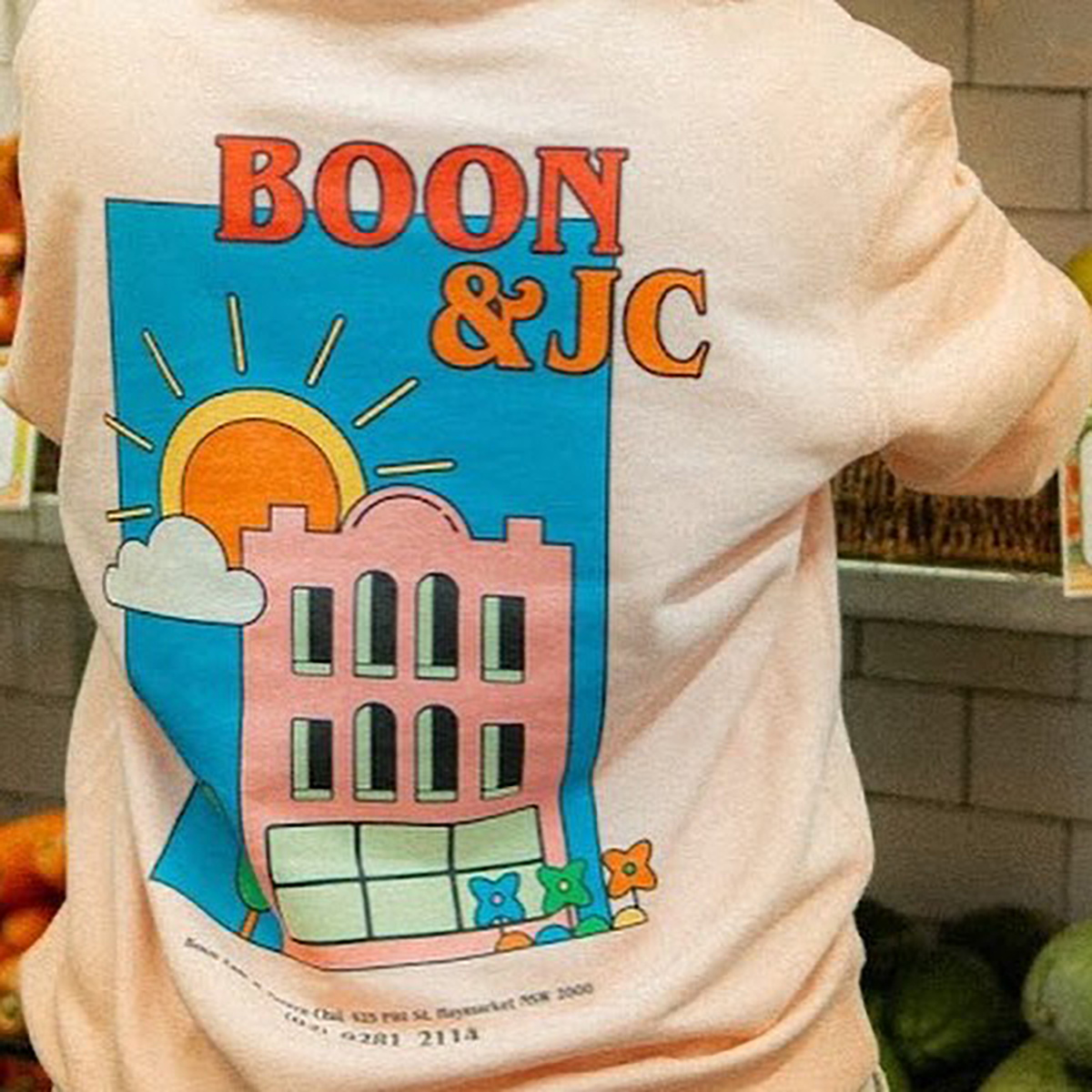 A printed peach tee worn in a Sydney grocer: a red BOON & JC wordmark over a blue, pink and yellow shopfront illustration